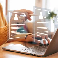 Henkilö käyttää kannettavaa tietokonetta pöydän ääressä ja selaa virtuaalisia tiedostoja, jotka näkyvät läpinäkyvinä kuvakkeina näytön edessä.