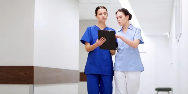 Kuvassa seisoo kaksi naishoitajaa sairaalan käytävällä ja tarkastelevat mustaa lehtiötä. 