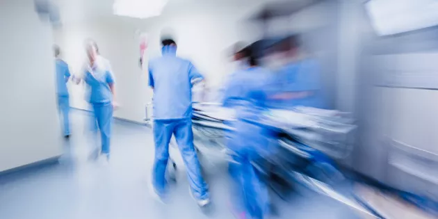 Kuvassa on siniasuisia hoitajia, jotka kiidättävät potilasta sairaalasängyssä eteenpäin sairaalan käytävällä.
