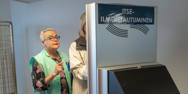 Itseilmoittautumisautomaatilla tuntematon asiakas, jota vaaleanvihreään vapaaehtoisten liiviin pukeutunut Thea Nurmi neuvoo.