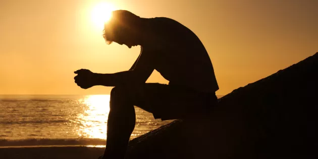 Kuvassa on miehen siluetti sivusta, joka istuu merenrannalla pääpainuksissa ja takaa paistaa aurinko.