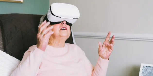Vanhempi nainen istuu tuolissa vr-lasit päässään ja kurottelee käsillä ylöspäin.