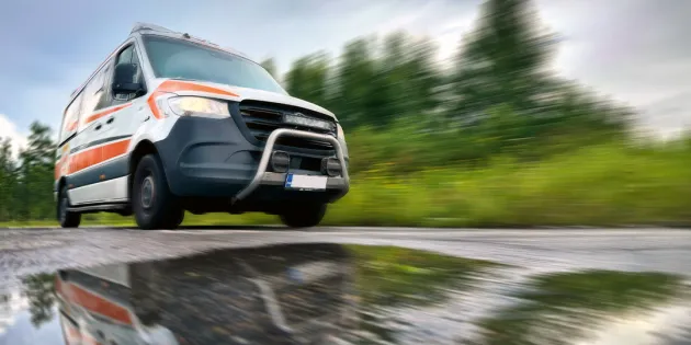 Ambulanssi ajaa kovaa vauhtia asfalttitietä, jonka päällä vesilammikko.