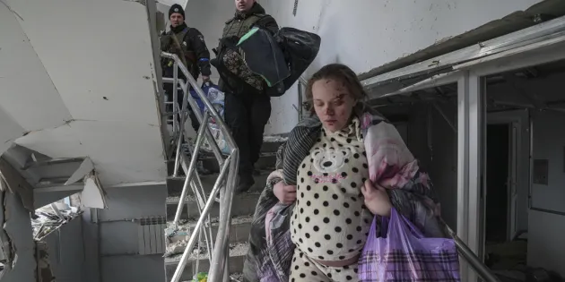 Raskaana oleva nainen, Mariana Vishegirskaya, laskeutuu portaita pitkin vaurioituneessa synnytyssairaalassa Mariupolissa, Ukrainassa. Hänen kasvoissaan on haavoja, ja hän kantaa peittoa ja muovikassia. Taustalla kaksi miestä seuraa häntä kantamuksineen. Sairaalarakennus on kärsinyt vakavia vaurioita