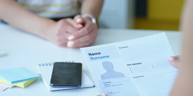 Henkilö istuu työhaastattelussa. Etualalla toinen henkilö pitää käsissään ansioluetteloa, jossa lukee &quot;Name Surname&quot;. Pöydällä on muistilehtiö, älypuhelin ja värikkäitä muistilappuja.