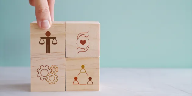 Uudistetuissa eettisissä ohjeissa käsitellään muun muassa rajallisten voimavarojen kohdentamista, teknologian roolia sekä ammattilaisten kokeman eettisen kuormituksen tunnistamista. Käsi asettelee puupaloikoita pinoon, jossa erilaisia symboleita.