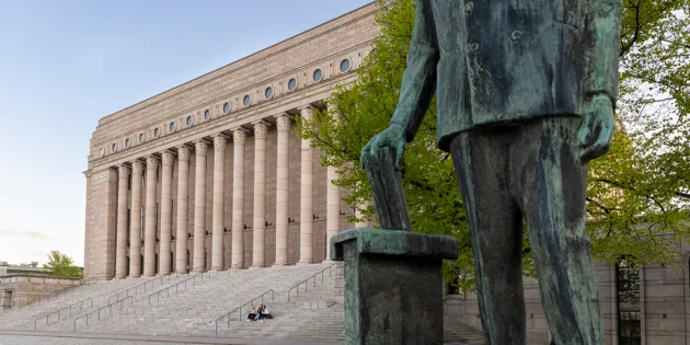 Eduskuntatalo Helsingissä kevätauringossa. Etualalla näkyy työväenjohtaja Yrjö Mäkelinin patsas, jonka hahmo pudottaa paperin vaaliuurnaan.