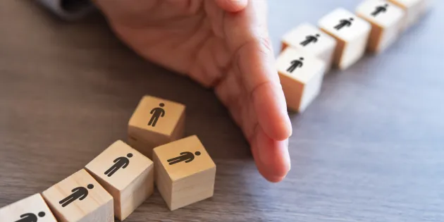 Laboratorioyhtiö Fimlab on aloittanut muutosneuvottelut, jotka voivat johtaa 30 henkilötyövuoden vähentämiseen. Käsi estää puisista palikoista muodostetun ihmisjonon etenemistä, yksi palikka on kaatunut muiden edessä.