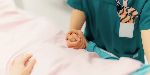Hoitaja pitää potilaan kädestä kiinni. Potilas makaa potilasvuoteella.
