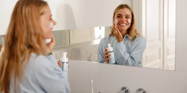 Nainen levittää voidetta kasvoilleen kylpyhuoneessa ja katsoo itseään samalla peilistä hymyillen.
