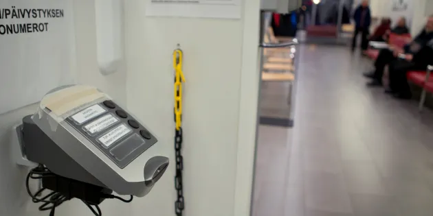 Odotustilassa vuoronumerolaite ja potilaita käytävän reunalla istumassa.