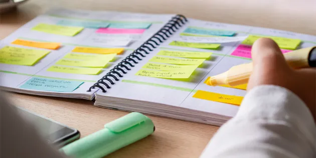 Henkilö kirjoittaa keltaisella korostuskynällä muistivihkoon, joka on täytetty värikkäillä post-it-lapuilla ja tehtävälistoilla. Vihko on avoinna pöydällä, jonka ääressä näkyy myös toinen korostuskynä ja älypuhelin.