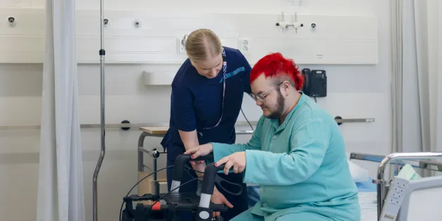 Hoitaja auttamassa potilasta nousemaan kävelytuen avulla ylös sairaalasängystä.