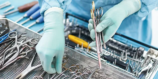 Hoitaja suojavarusteissa järjestelee instrumentteja leikkaussalissa valmiiksi toimenpiteeseen, Kuvassa näkyvät kädet ja instrumentteja.