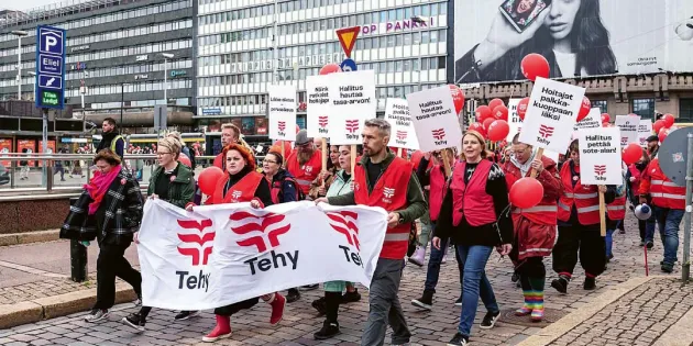  Tehyn jäseniä marssii kadulla Helsingissä punaisiin liiveihin pukeutuneina ja kantaen banderollia, jossa lukee &quot;Tehy&quot;. Useilla on kylttejä, joissa vaaditaan parempaa palkkaa ja arvostusta hoitajille. Taustalla näkyy Elielinaukio, Yliopiston Apteekki, mainostauluja ja Helsingin keskustan rakennuksia.