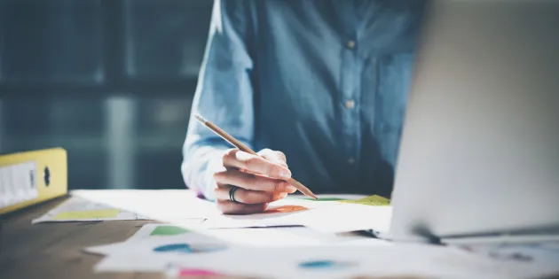 Henkilö laatimassa tietokoneella suunnitelmaa ja hahmottelee samalla kynällä paperille.