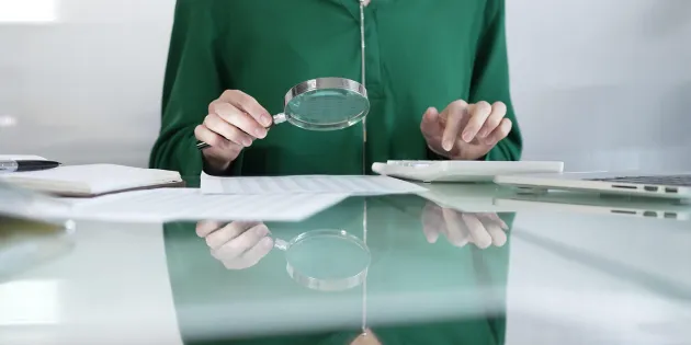 Ihminen istuu pöydän ääressä ja tutkii suurennuslasilla papereita. 