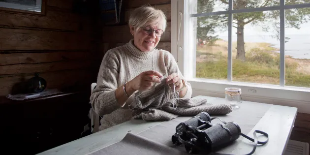Tuovi Anttila neuloo mökillä pöydän ääressä ikkunan edessä.