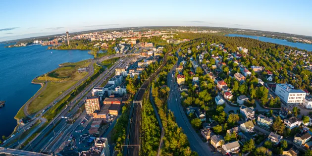 Ilmakuva Tampereen kaupungista kesäiltana, etualalla rautatie ja asuinalue värikkäine taloineen, vasemmalla suuri vesialue ja satama-alue, taustalla keskustan rakennuksia ja näkötorni, oikealla toinen vesialue ja vihreitä metsäalueita.