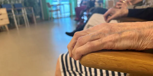 Vanhuksen käsi lepää tuolin selkänojalla. Vanhukset istuvat ringissä palvelutalon yhteisessä tilassa.