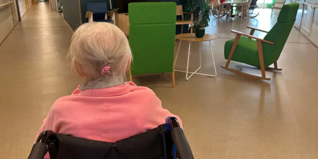 Vanhus istuu pyörätuolissa selin kameraan hoivakodin aulatilassa.