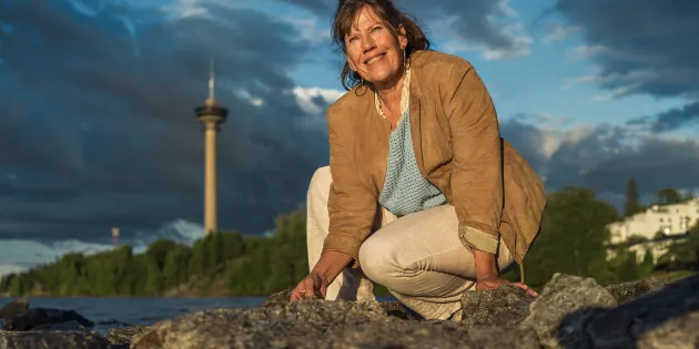 Ulla Ollikkala kyykyssä rantakivikossa, taustalla Tampereen Näsinneula.