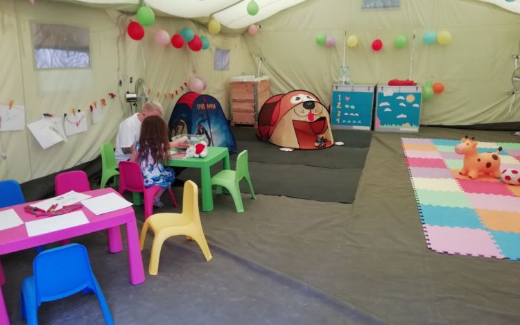 Värikkäitä lasten pöytiä, tuoleja ja leikkivälineitä ison teltan nurkassa.