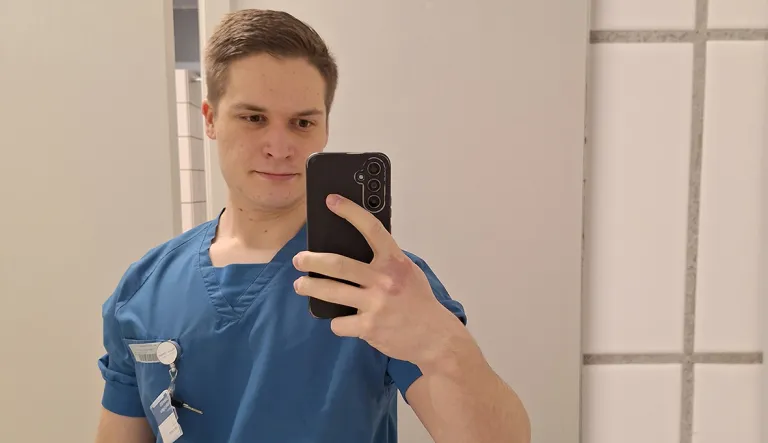 Arttu Mikkola kuvaa itseään kännykällä peilin kautta. Päällään hänellä on sinivihreä työasu.