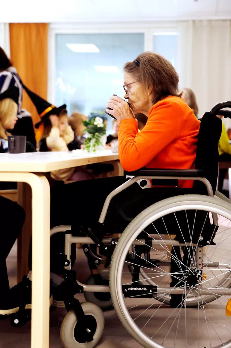 Pyörätuolissa istuva vanha rouva siemailee kahvia.