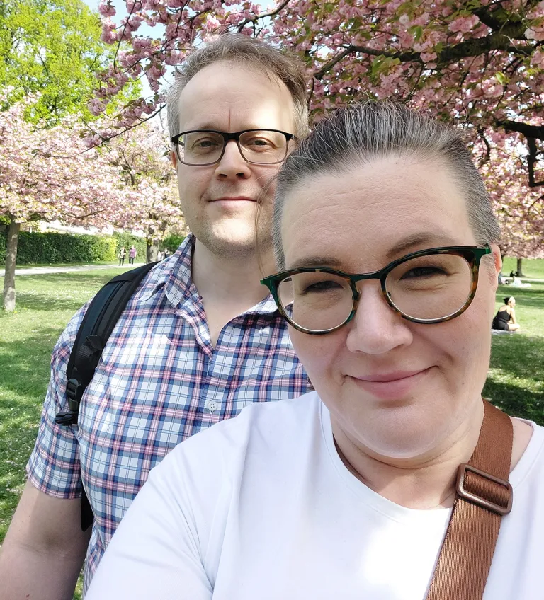Anne ja Jyri Meriläinen puistossa kirsikkapuiden kukkiessa taustalla.