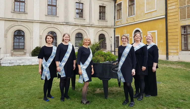 Kuusi naista seisoo nurmikolla historiallisen rakennuksen edustalla. Heillä on yllään mustat mekot ja olkapäiden yli kulkevat sinivalkoruudulliset nauhat. He hymyilevät kameralle ja seisovat vanhan mustan pianon edessä, joka on asetettu ulkotilaan ja koristeltu vihreillä kasveilla. Taustalla näkyy vanhoja ikkunoita ja keltainen sekä beige julkisivu.
