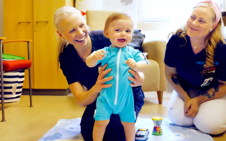 Fysioterapeutti Taru Palsa pitelee pystyssä yhdenksänkuista Rafael-vauvaa. Tutkimushoitaja Jonna Salama istuu vieressä.