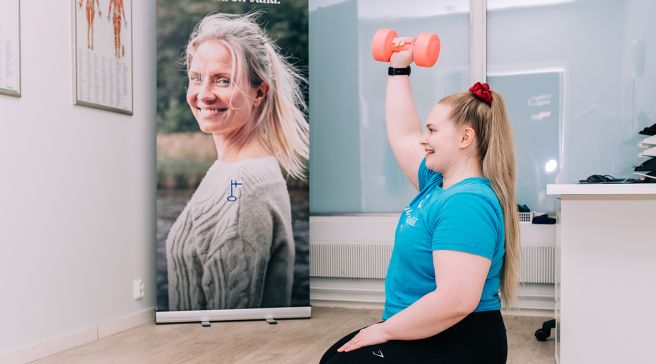 Fysioterapeutti Kaisa Jakonen kuinka harjoitellaan käsipainoilla. Istuu polvillaan lattialla sinisen jumppamaton päällä.