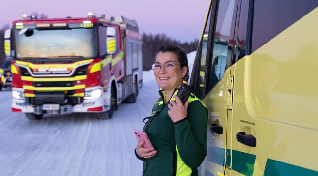 Ensihoitaja Niina Leskinen poseeraa talvisella tiellä nojaten ambulanssiin kännykkä ja radio kädessään.