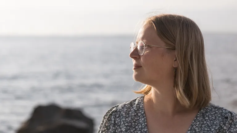 Sairaanhoitaja Mirva Virtanen seisoo rannalla kesämekossa ja katsoo horisonttiin vakavana.