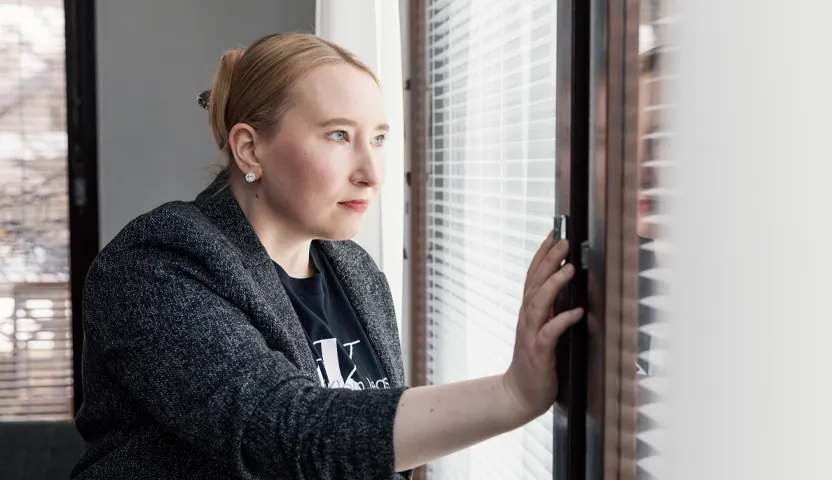 Sairaanhoitaja Annamari Viljanen katsoo ikkunasta ulos sälekaihtimien läpi.
