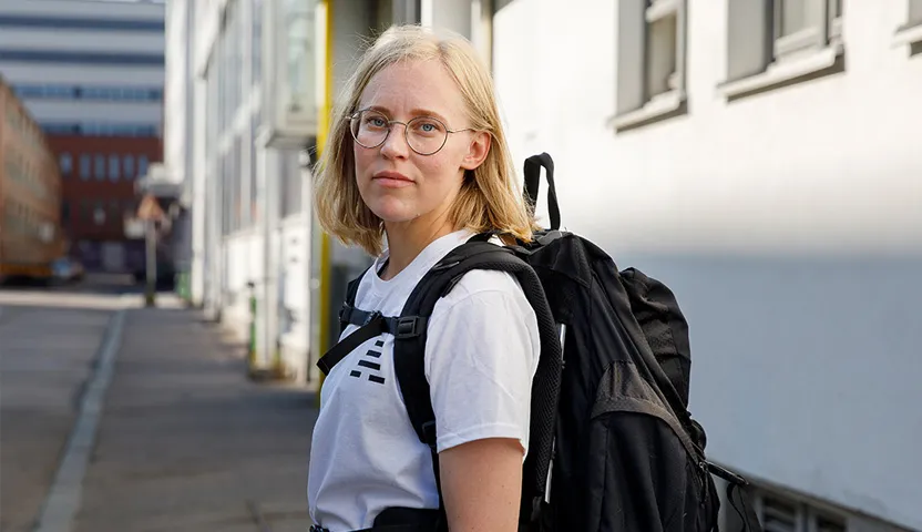 Sairaanhoitaja Maija Vanhala seisoo kadulla selässään suuri musta reppu. Hänellä on vaaleat hiukset, silmälasit ja valkoinen t-paita.