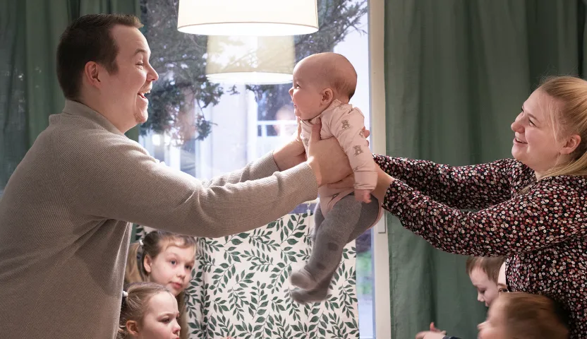 Rosenbergin perhe keittiön pöydän ääressä. Äiti ojentaa nuorimmaista lasta isälle pöydän yli..