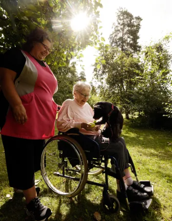 Mari Hyökki seisoo pyörätuolin vieressä. Pyörätuolissa istuu Aili Erola Velma-koira sylissään.