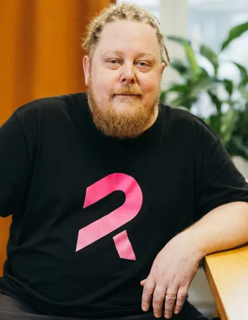 Rami Oksanen, katsoo kameraan ja nojaa pöytään. Musta paita päällä, jossa pinkki kuvio.