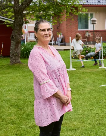 Pirkko Peltomaa kotinsa takapihalla. Taustalla lapsenlapset keppihevosen selässä.