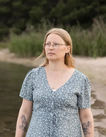 Sairaanhoitaja Mirva Virtanen seisoo rannalla kesämekossa ja katsoo horisonttiin vakavana.