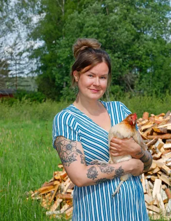 Julia Lehtiä seisoo pihallaan kana sylissään. Takana kasa halkoja ja vihreää nurmea.